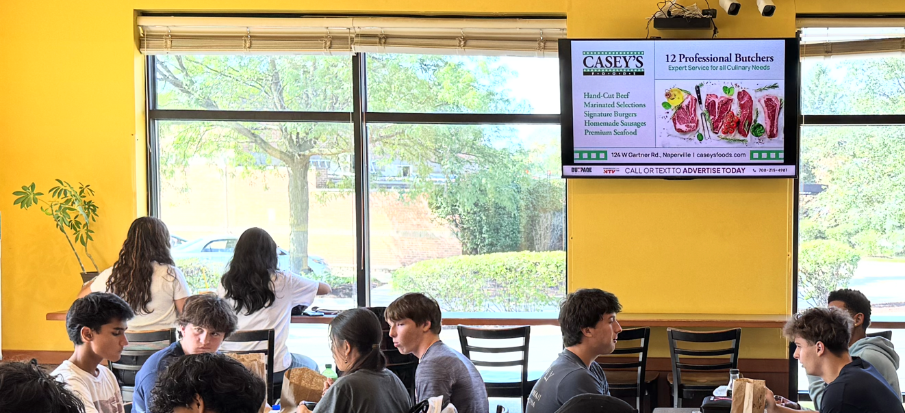 DDM digital advertising screen inside Big Apple Bagels in Aurora, with customers in the venue