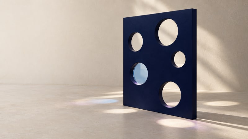 Matte navy panel with five circular openings on a sunlit off-white floor; one opening holds a pale-blue disc.