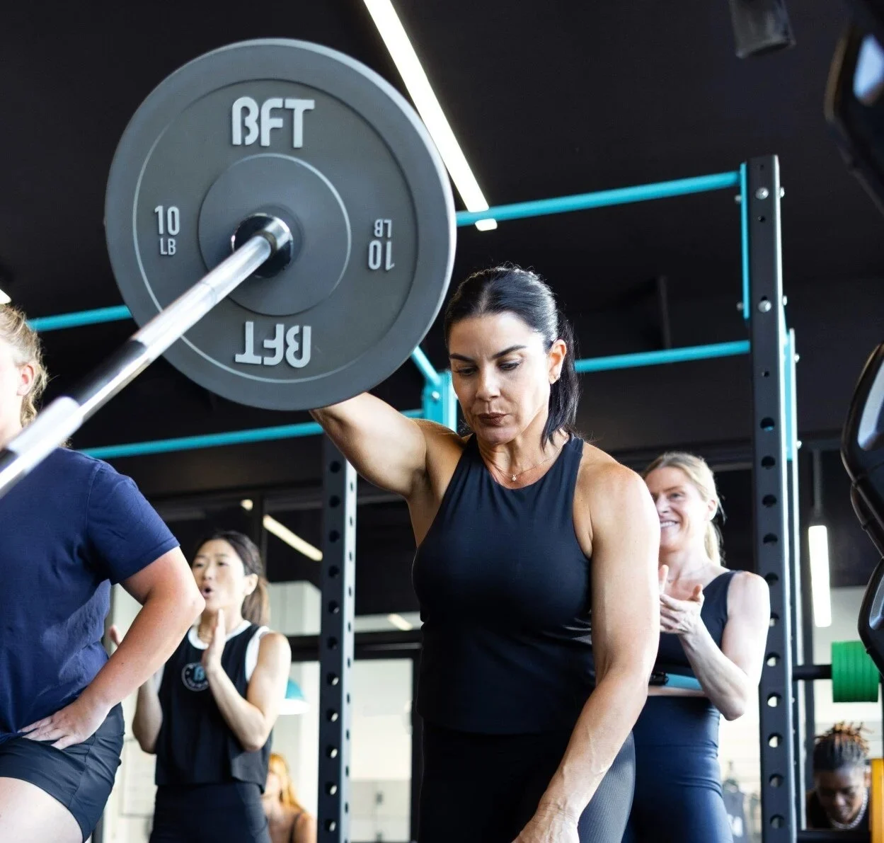 Woman lifting a barbell in a BFT group strength class, with a BFT weight plate in the foreground