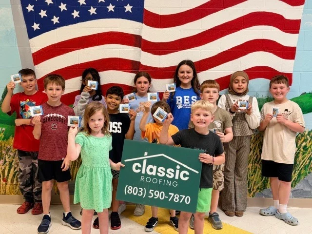 Children holding a Classic Roofing sign in front of an American flag backdrop